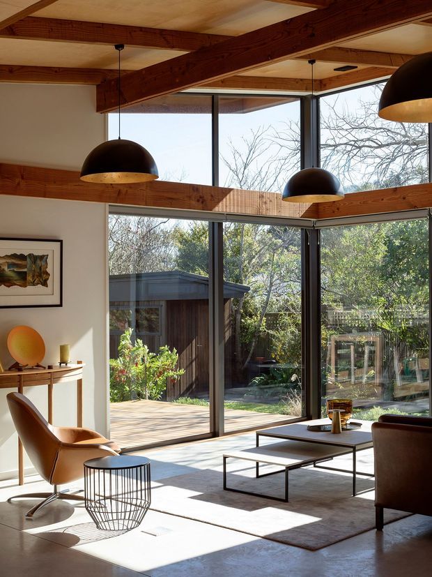 A sunlit mid-century-inspired living room with exposed timber ceiling beams, expansive floor-to-ceiling glazing, and direct views to a leafy garden courtyard. The room contains sculptural pendant lights, a low rectangular coffee table, a leather lounge chair, a sofa, a wire-frame side table, a console, and a neutral rug. Dark-framed glazing and warm timber details create a strong indoor–outdoor connection.