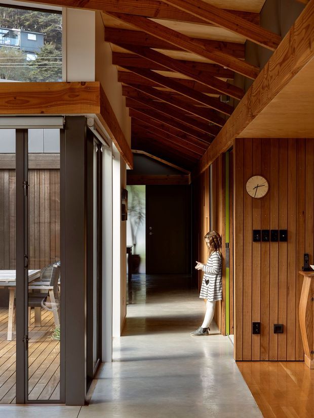 A long, light-filled residential hallway with a dramatic exposed timber pitched roof and richly stained timber wall panelling. Full-height glazed doors open to a timber deck and outdoor seating on the left, while built-in timber joinery and dark electrical fittings line the right wall. Polished grey concrete flooring transitions to warm timber flooring, creating a strong contrast between materials. A child stands midway down the corridor, with further rooms visible beyond.