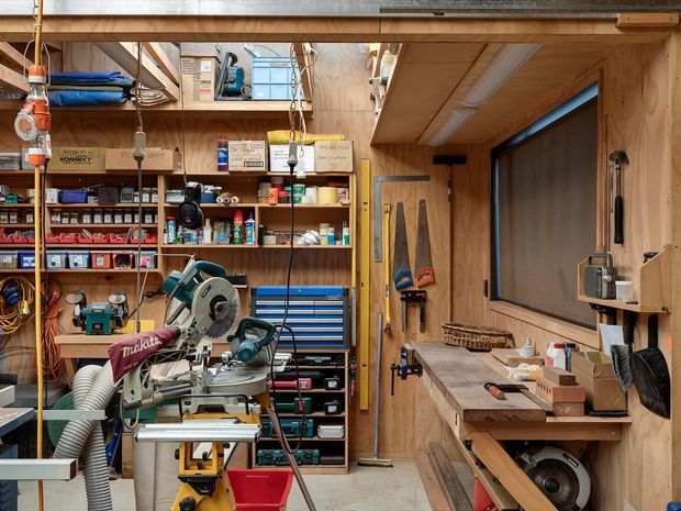 A compact, highly organised woodworking workshop with extensive open shelving, boxed supplies, hand tools, power tools, a mitre saw, workbenches, clamps, and a large dust-extraction hose. The room is lined with warm timber plywood on the walls and ceiling, with built-in storage, a timber workbench, a large window, and bright task lighting. The overall appearance is practical, workshop-oriented, and richly detailed.