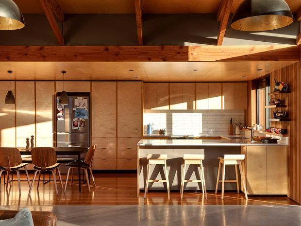Warm, sunlit open-plan kitchen and dining area with extensive light timber cabinetry and wall panelling, a central island with three stools, and a dining table with wooden chairs. Exposed timber roof beams and a dark pitched ceiling add architectural character, while pendant and dome lights provide layered illumination. A large window brings strong afternoon light across the white tiled backsplash and polished timber and concrete floors.