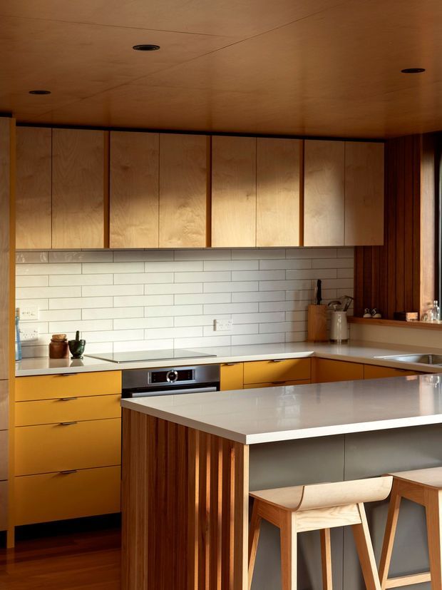 Warm, compact contemporary kitchen with extensive timber cabinetry and ceiling, a white horizontal subway-tile splashback, mustard-yellow lower drawers, and a large pale stone island with integrated seating. Vertical dark timber battens wrap the island end and wall, while recessed ceiling lights and streamlined appliances create a refined, cohesive fit-out.
