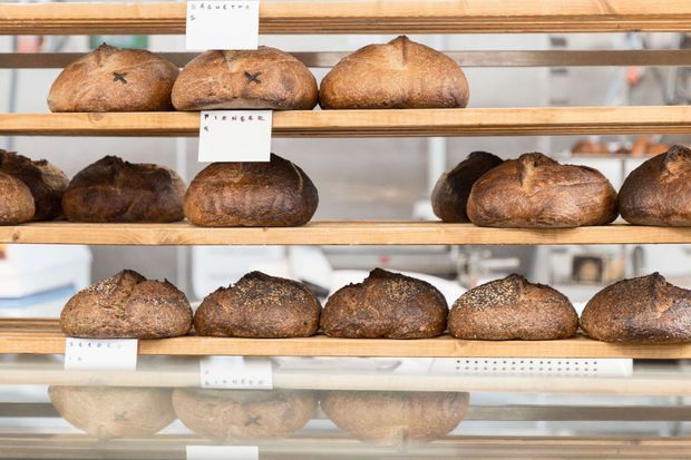 Close-up of assorted artisan loaves displayed on several horizontal wooden bakery shelves, with small handwritten labels and reflections in the glass or polished surface below. The image focuses on bread presentation in a bakery or commercial food setting.