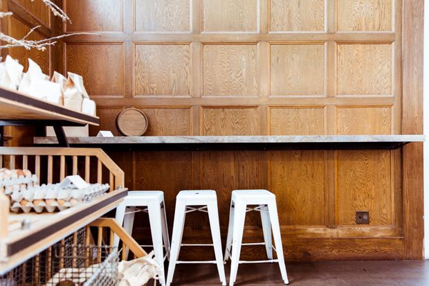 A warmly lit commercial-style counter or café space featuring a full-height dark timber wall with traditional rectangular panel moulding, a long pale stone counter, and three white metal stools. Timber display shelving with trays and packaged goods occupies the left foreground, while a dark timber floor and visible power outlet complete the scene.