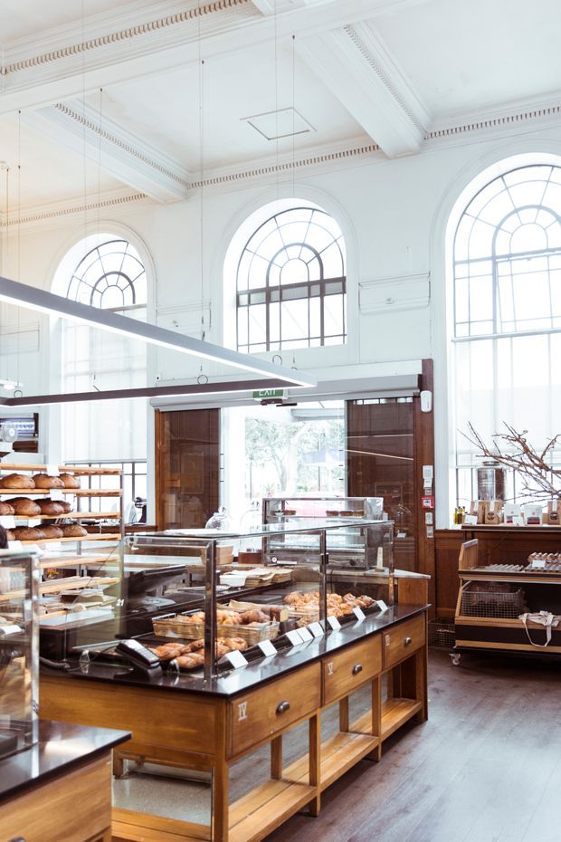 Bright, high-ceilinged bakery or café interior in a historic-style commercial space. The room features tall arched multi-pane windows, ornate white ceiling cornices and decorative mouldings, exposed suspended linear lighting, dark timber-framed glazed entry doors, and multiple polished timber-and-glass pastry display counters filled with baked goods. The fit-out combines heritage architectural detailing with refined contemporary retail fixtures.