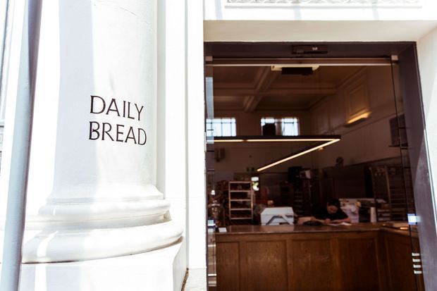 Exterior-facing view into a bakery storefront, with a white classical-style column bearing the words “DAILY BREAD” beside a dark-framed glazed entrance. Inside, warm timber cabinetry and counters, shelving, pendant lighting, and a high coffered ceiling are visible.