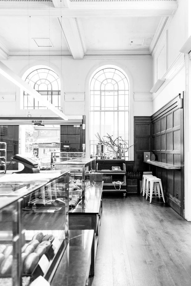 Black-and-white photograph of a refined bakery or café interior in a tall heritage-style space. Glass pastry display counters occupy the foreground, while the rear wall features dark timber raised-panel wainscoting, a narrow stone or composite counter with white stools, and a mobile shelving unit. Large arched multi-pane windows, ornate ceiling mouldings, decorative cornices, and exposed ceiling beams create a bright, formal architectural character.