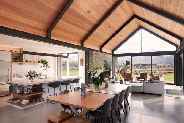 A spacious open-plan contemporary home interior combining a kitchen, dining area and living room beneath a dramatic exposed timber vaulted ceiling. Black structural beams frame the warm timber ceiling, while extensive floor-to-ceiling glazing opens the living area to a rural landscape. The kitchen features a pale island with bar seating, open shelving and integrated cabinetry; the dining area has a large timber table with dark upholstered chairs, and the living zone includes a light sofa and lounge chairs.