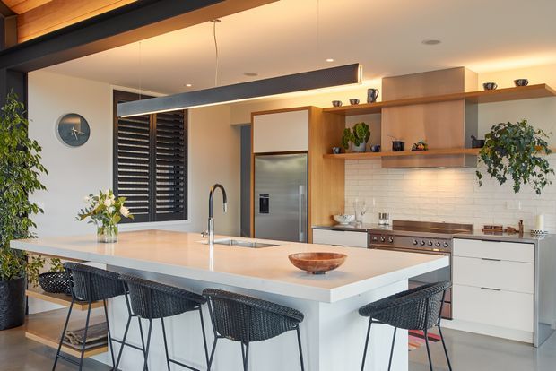 A warm, contemporary open-plan kitchen with a large pale island and integrated sink, four woven bar stools, stainless-steel appliances, white cabinetry, timber open shelving, and a white tiled splashback. Black-framed glazing with external louvres, indoor plants, and a long linear pendant light add contrast and texture.