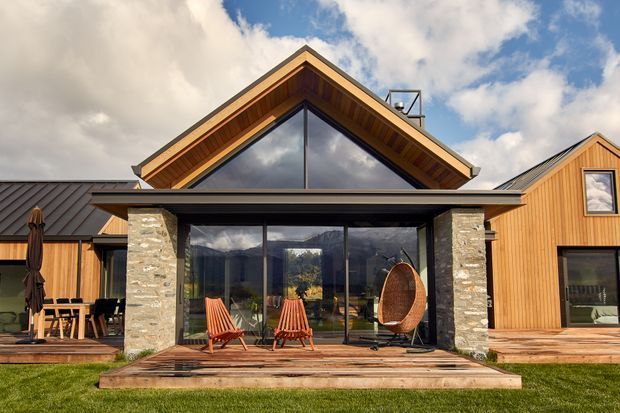 Exterior view of a contemporary timber-clad, gable-roofed house with a prominent glazed gable, dark metal roofing, stone-clad structural piers, and timber decking. The central outdoor entertaining area includes seating and a hanging chair, with landscaped lawn in front and mountainous scenery reflected in the glazing.