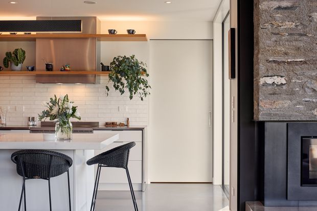 A refined contemporary kitchen and open-plan living area. The kitchen features pale cabinetry, a white subway-tile splashback, a light stone island benchtop, warm timber open shelving, integrated appliances, and black woven bar stools. A black fireplace with a textured stone surround is visible on the right, alongside full-height glazing and a light polished floor.