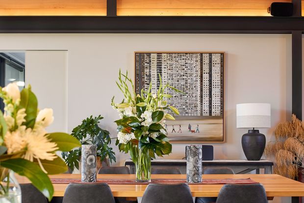 Warm, refined dining room with a long timber dining table, dark upholstered dining chairs, and a sideboard beneath framed artwork. A large floral arrangement forms the centrepiece, with decorative plants, lamps, and sculptural accessories. The room has a neutral plaster wall, dark structural beams, and warm concealed lighting.