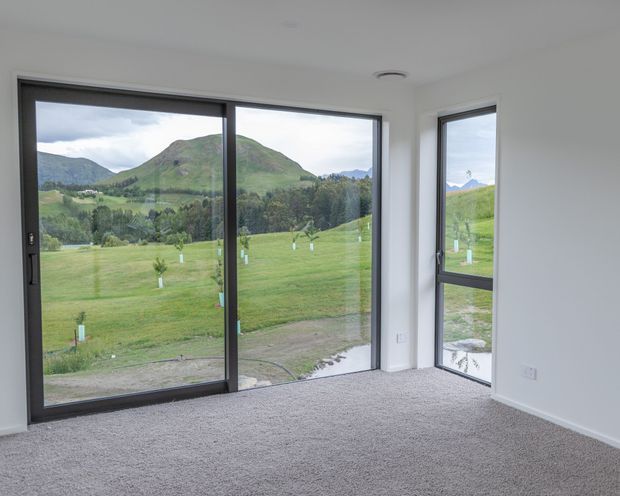 Empty, bright interior room with grey carpet, white walls and ceiling, and large dark-framed floor-to-ceiling windows and sliding glass doors overlooking a green rural valley and mountains. The room has a simple contemporary shell with abundant natural light and no furnishings.