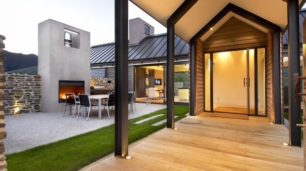 Contemporary residential courtyard and entry viewed from a timber deck. The composition features a covered outdoor walkway with black structural posts, a warm timber-clad entry volume with glazed double doors, a corten- or dark-metal-roofed pavilion, a concrete outdoor fireplace and dining area, stone walls, gravel paving, and narrow lawn strips. Mountainous terrain is visible in the background.