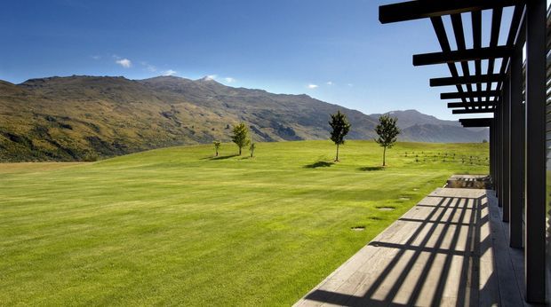 A wide exterior view from a modern building looking across a manicured lawn toward rugged mountainous terrain. A timber deck and strongly projecting dark pergola beams run along the right side, casting long linear shadows across the decking. The scene is open, sunny, and strongly connected to the surrounding landscape.