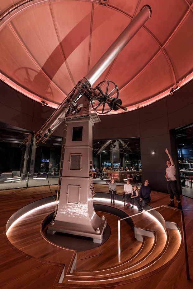 A dramatic interior of a public observatory or planetarium featuring a large historic telescope mounted beneath a domed, segmented ceiling. The telescope stands on a substantial central pedestal surrounded by concentric stepped timber platforms with integrated lighting and seating. Dark glazed walls reveal a café or exhibition area beyond, while visitors gather around the exhibit. The space is illuminated with warm red and amber lighting, creating a theatrical museum-like atmosphere.