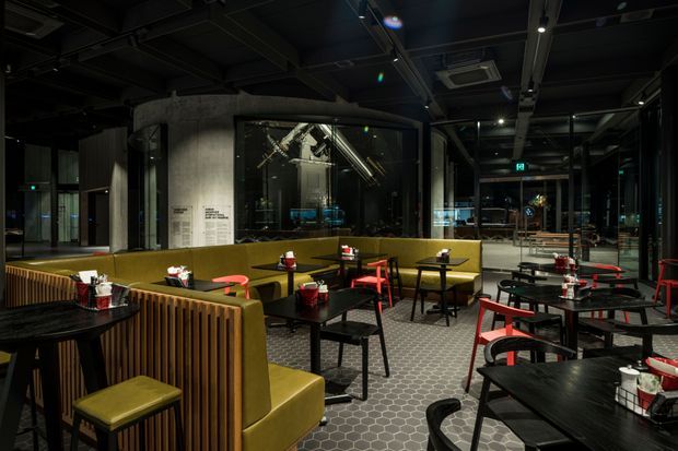A dark, sophisticated commercial dining or café interior with olive-green upholstered booth seating, red and black chairs, small black tables, and a patterned hexagonal floor. The space features exposed dark ceiling services, concrete columns, extensive black-framed glazing, and a modern industrial fit-out with warm accent lighting.