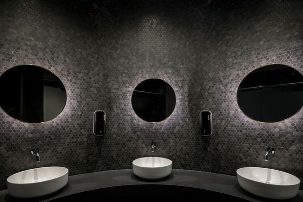 A dramatic, symmetrical bathroom vanity featuring three white vessel basins on a dark curved countertop. The walls are fully clad in small dark grey mosaic tiles, with three circular mirrors, black wall-mounted soap dispensers, and black tapware. The space has a moody, refined, contemporary appearance with strong repetition and low lighting.