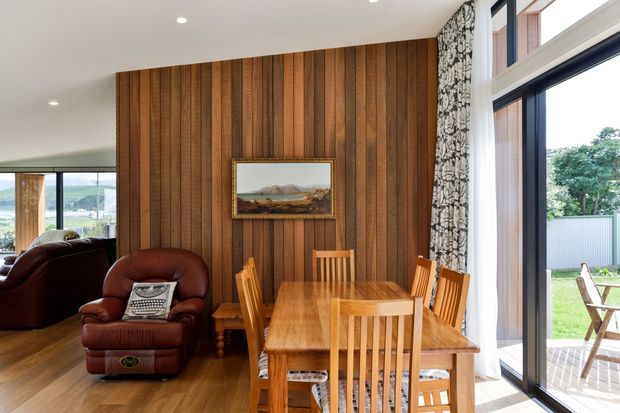 Warm, timber-lined dining area with a solid wooden dining table and matching chairs, opening toward a bright living space and glazed outdoor terrace. The feature wall has vertical dark timber boards, framed artwork, and patterned curtains beside large windows and a sliding glass door. A leather armchair and lounge area are visible to the left.