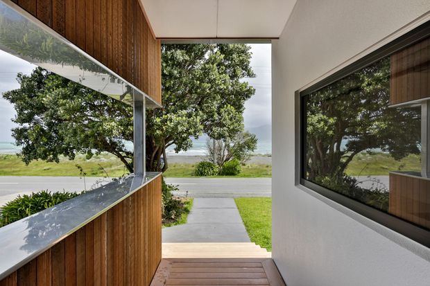 A contemporary covered entryway or passage framed by warm vertical timber cladding, white rendered walls, and large dark-framed glazed openings. Reflections extend the view of the surrounding trees and lawn, with a concrete path leading toward a quiet coastal or waterfront landscape. Timber decking and a clean linear roof soffit reinforce the modern architectural character.