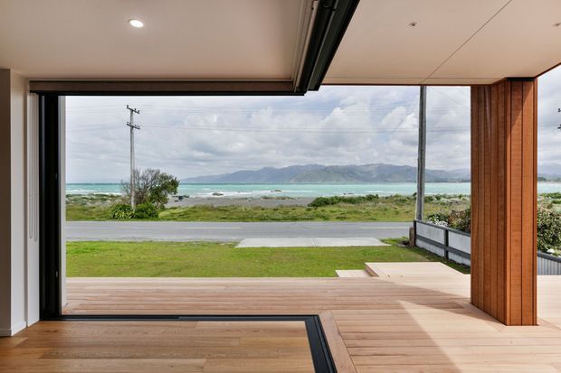 A contemporary indoor-outdoor living space with wide-open glazed sliding doors framing a coastal view of turquoise water and mountains. The room features warm timber flooring, a timber-clad structural column, a covered deck, and a clean white ceiling with recessed lighting.