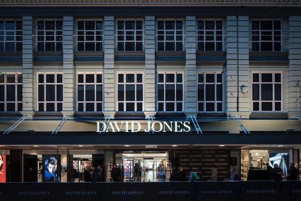 Street-level view of a historic multi-storey commercial department-store facade. The upper facade features repeated tall multi-pane windows, pale horizontal masonry or rendered bands, projecting pilasters, and a traditional cornice. The ground floor is a contemporary retail frontage with large glazed shopfronts, illuminated interiors, signage reading “DAVID JONES,” and temporary event barriers along the footpath.