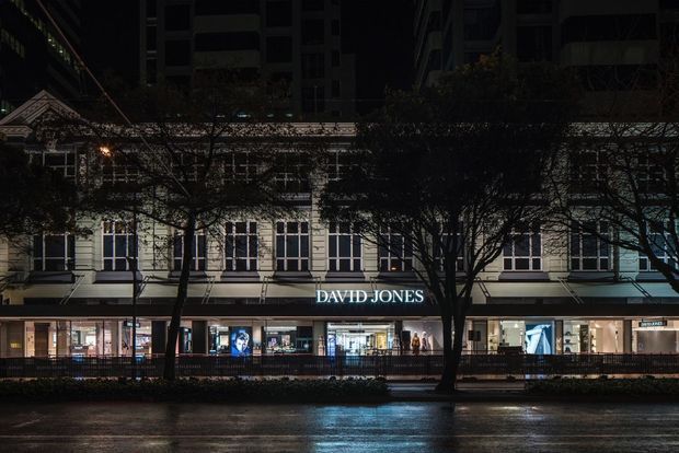 Night-time street-level view of a multi-storey commercial retail building, with the illuminated David Jones department store frontage, repetitive tall windows, decorative upper facade detailing, street trees, and wet pavement reflecting the storefront lighting.