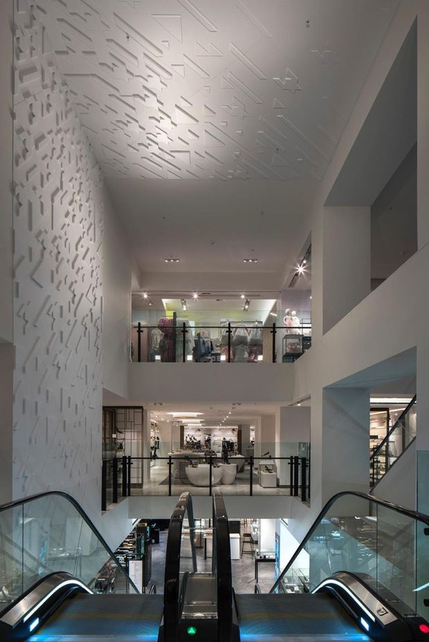 Interior view of a multi-level contemporary retail or commercial space, photographed from an escalator. The atrium features multiple open levels, glass balustrades, white structural walls, recessed and track lighting, and a highly distinctive three-dimensional geometric relief pattern across the left wall and ceiling. Clothing displays and retail fixtures are visible on the upper and lower floors.