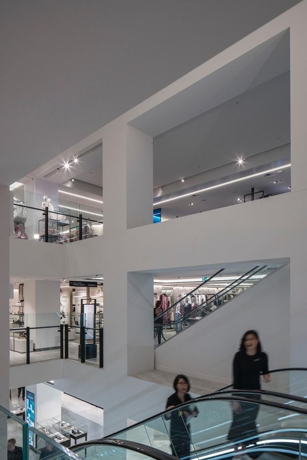 Interior view of a multi-level commercial retail space, likely a department store or shopping centre, with large white structural openings, multiple floor levels, glass balustrades, escalators, recessed spotlights, linear ceiling lighting, and visible retail displays. The image is photographed from an escalator and includes shoppers moving through the space.
