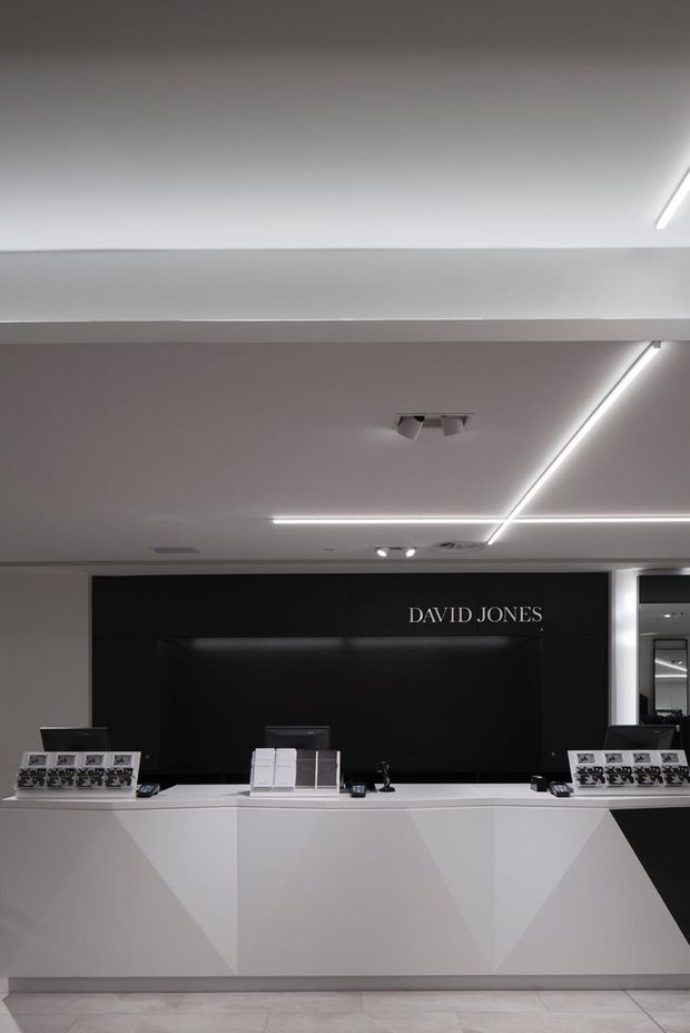 A modern retail or department-store service counter for David Jones, featuring a long white geometric reception desk, a black back wall with illuminated branding, integrated display stands, and linear LED ceiling lighting. The monochrome fit-out has a clean, minimalist commercial aesthetic with pale tiled flooring and strong black-and-white contrast.