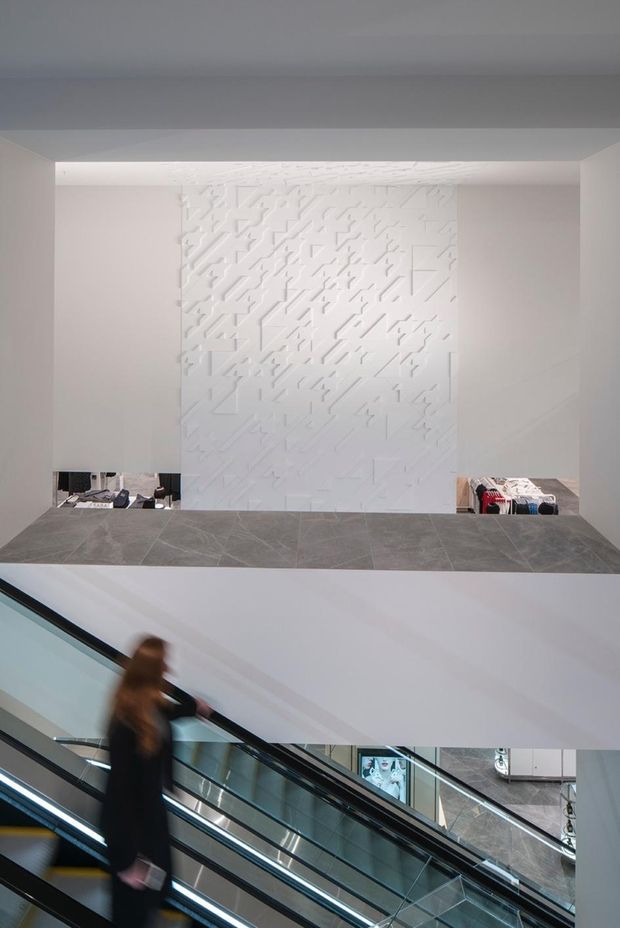A bright, minimalist commercial interior viewed across an escalator void. The scene features white architectural surfaces, a large textured geometric feature wall, grey stone-look horizontal ledges, extensive glass balustrades, and multiple escalators. Clothing displays visible through openings suggest a retail shopping environment.