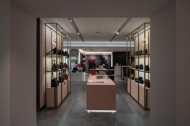 Interior view of a high-end retail showroom, framed by a dark doorway and centered on a pale blush display island. Symmetrical illuminated brass-toned shelving along both sides presents handbags and accessories, with additional merchandise displays toward the rear. The space has polished dark stone-look flooring, a white recessed ceiling with integrated spotlights and linear lighting, and a refined, gallery-like layout.