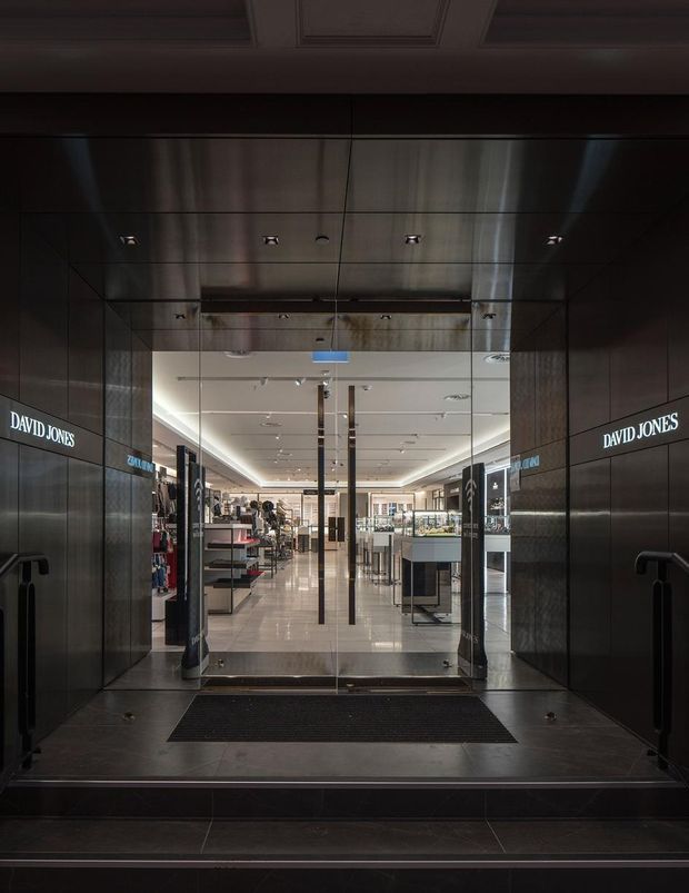 A dark, polished retail-store entrance leading into a bright department-store interior. The entry vestibule is clad in reflective dark panels with recessed downlights, glass double doors, black pull handles, and a black entrance mat. Illuminated David Jones signage appears on both side walls, while retail counters and displays are visible beyond the doors.