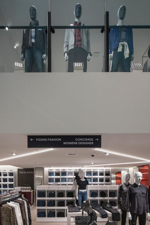 Interior view of a multi-level department store fashion area, with mannequins displaying clothing on an upper mezzanine and sales-floor racks, shelving, counters, signage, and display fixtures below. The space has a clean, contemporary retail fit-out with recessed and linear ceiling lighting, glass balustrading, neutral walls, and dark display elements.