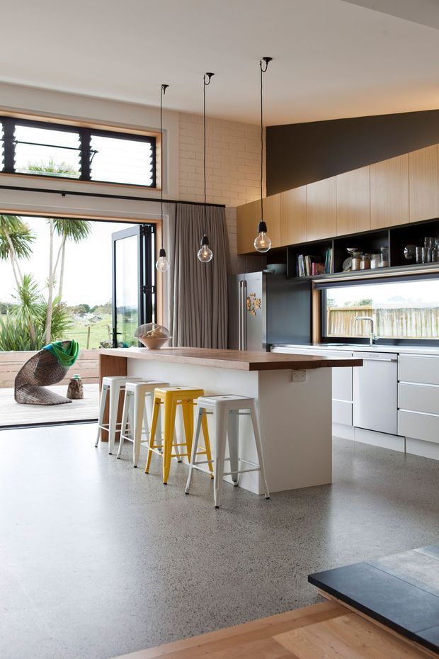Open-plan contemporary kitchen with a large island and integrated seating, featuring a polished concrete floor, white cabinetry, dark timber-toned overhead joinery, exposed white brick, pendant lights, and extensive indoor-outdoor glazing opening to a deck and garden.