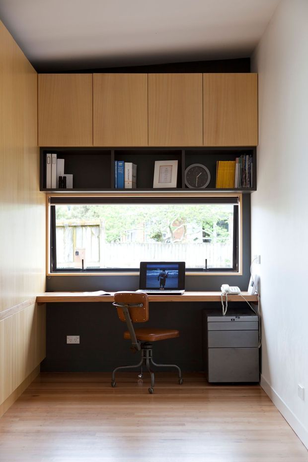 Compact built-in home office nook with a timber desk spanning beneath a large window. The space features warm timber cabinetry and flooring, dark shelving and wall backing, a vintage-style swivel office chair, filing drawers, a computer, and neatly arranged books and decor. Natural light enters through the window, creating a warm, functional workspace.