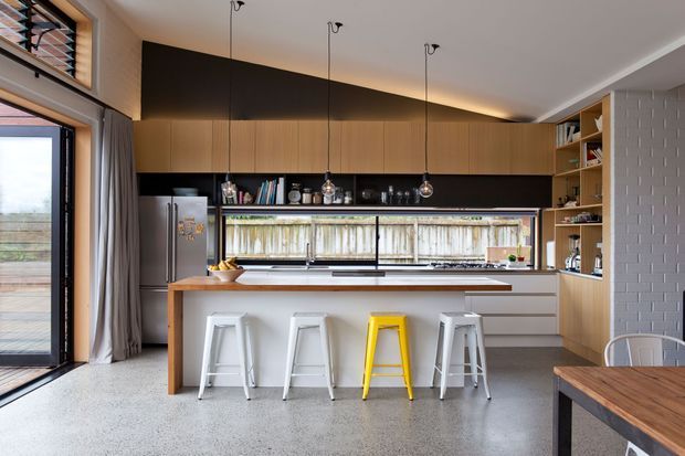Contemporary open-plan kitchen with a large timber-fronted island, integrated dark cabinetry, stainless-steel appliances, white bar stools with one bright yellow accent stool, pendant lights, open shelving, and a terrazzo-like grey floor. Full-height glazing and a timber-fenced outlook provide natural light, while a textured white tiled wall adds detail.