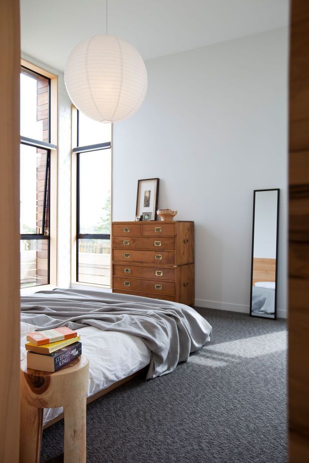 A bright, contemporary bedroom with a low platform bed dressed in rumpled grey bedding, a warm timber bedside stool, and a vintage timber chest of drawers. Tall black-framed windows bring in abundant natural light, while a large white paper pendant hangs from the ceiling. A full-length black-framed mirror leans against the wall, reflecting part of the bed. The room has white walls, grey carpet, and a calm, understated palette.