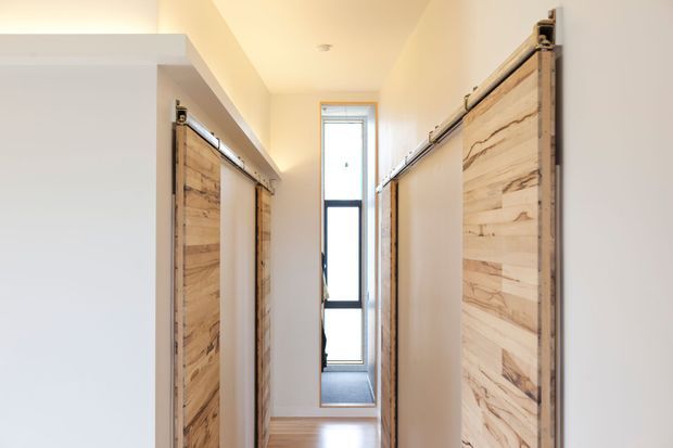 A narrow, bright interior passage lined on both sides with sliding barn-style doors featuring prominent natural timber grain and exposed metal hardware. White walls and ceiling, pale timber flooring, and a tall glazed door or window at the far end create a clean, warm corridor.