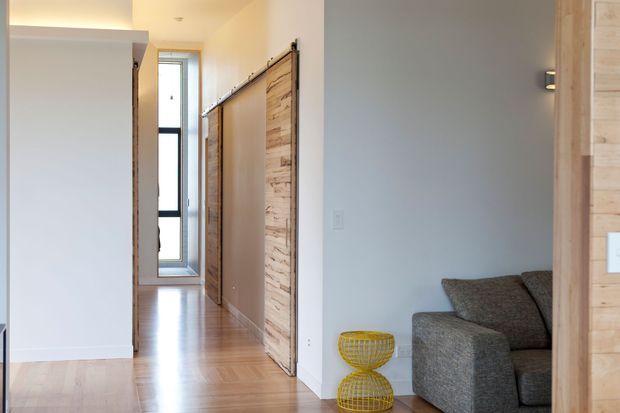 Bright contemporary interior showing a hallway with warm timber flooring, white walls, a narrow vertical window, and distinctive timber-clad sliding or barn-style doors. A grey upholstered sofa is partially visible in an adjoining living area, with a small yellow wire side table adding an accent.