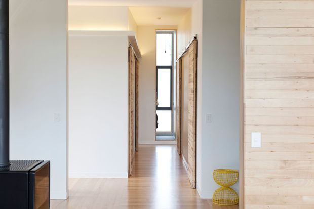 Bright contemporary hallway or entry passage with pale timber flooring, white walls, exposed light timber wardrobe or storage doors, and a tall narrow window at the far end. A light timber wall panel and small decorative wire stools add warmth to the minimalist interior.