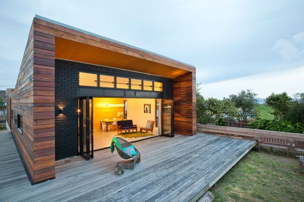 Contemporary timber-clad pavilion-like house extension with a deep recessed outdoor opening, black masonry wall, warm timber soffit, and extensive weathered timber decking. Large glazed folding or sliding doors open the interior living area to the deck, with louvred clerestory windows above and a rural landscape beyond.