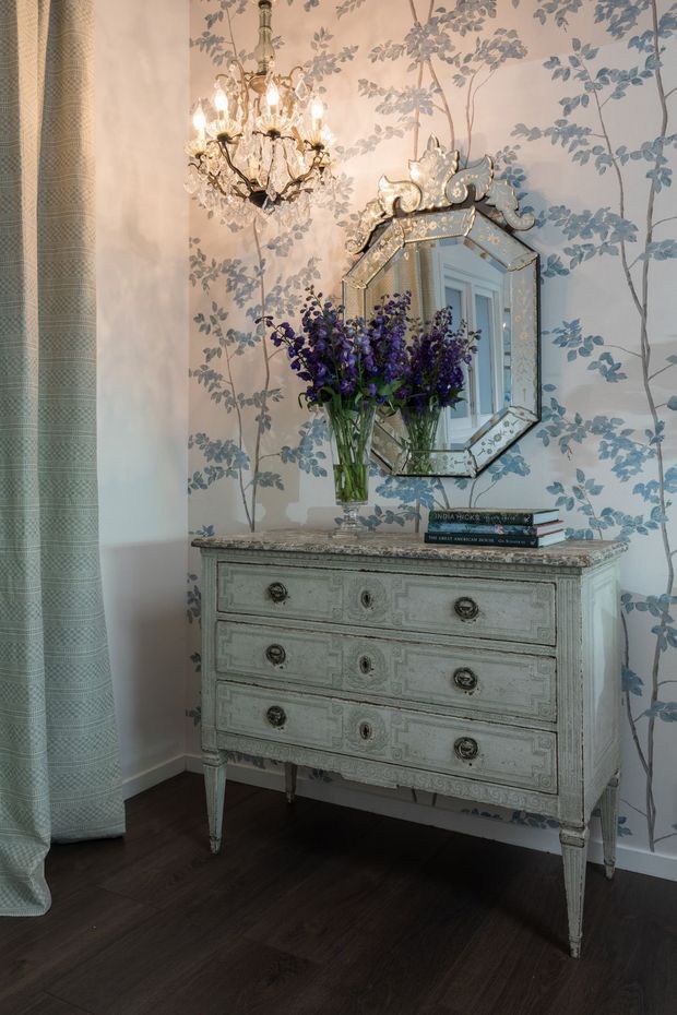 A refined traditional interior vignette featuring an ornate pale blue-grey antique-style chest of drawers against blue botanical wallpaper. An elaborate faceted mirror, crystal chandelier, floral arrangement, patterned curtain, and dark timber flooring create a decorative, layered and elegant atmosphere.
