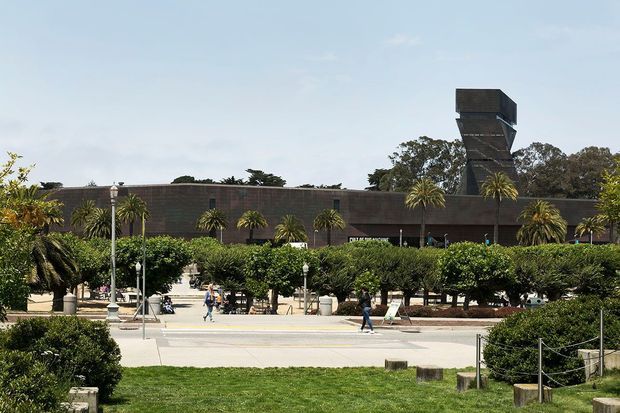 A low, dark brick institutional building set behind a landscaped public forecourt, with rows of manicured hedges, palm trees, lawn, paved walkways, and people walking through the space. A prominent angular tower rises above the flat roofline, creating a strong geometric architectural feature.