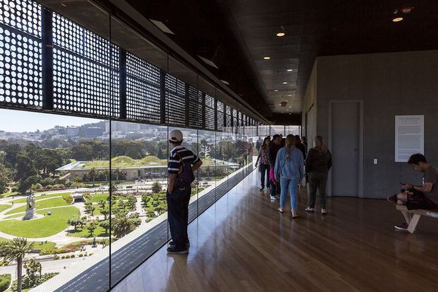 A long, elevated interior gallery or circulation corridor with a polished timber floor, dark ceiling, concrete-toned walls, and extensive floor-to-ceiling glazing. Perforated metal screening runs along the upper edge of the glazed facade, filtering daylight and creating a strong geometric pattern. Visitors stand and sit along the corridor, while landscaped grounds and a cityscape are visible beyond the glass.
