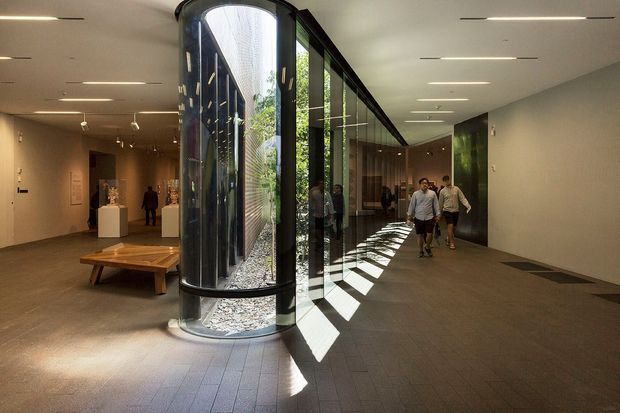 Contemporary gallery or museum-like interior with a long circulation corridor, dark tiled flooring, recessed linear ceiling lights, white walls, and extensive floor-to-ceiling glazing. A distinctive curved glazed display enclosure contains a planted feature, while strong sunlight creates geometric shadows across the floor. Visitors and sculptural display plinths are visible in the background.