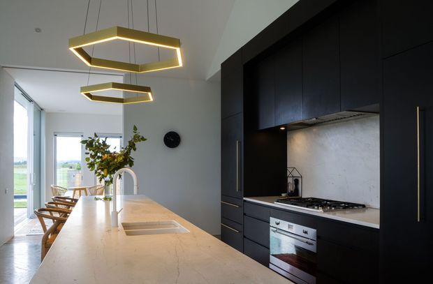 Contemporary kitchen with a long pale stone island featuring an integrated sink and arched metal tap, dark floor-to-ceiling cabinetry, a matching pale textured backsplash, and a gas cooktop. A timber dining area and large windows sit beyond the island, while geometric pendant lights add a warm accent.