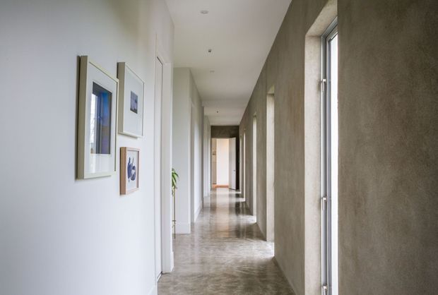 A long, narrow contemporary hallway with polished concrete flooring, white walls, framed artwork, recessed ceiling lights, and tall narrow windows set into a textured grey wall. The corridor leads toward a brighter room at the far end and has a clean, minimalist architectural character.