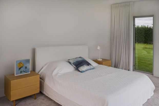 A calm, minimalist bedroom with a neatly made double bed, white upholstered headboard, pale bedding, light grey walls, and full-height curtains beside a tall window overlooking greenery. Matching light timber bedside furniture and a simple bedside lamp complete the understated interior.