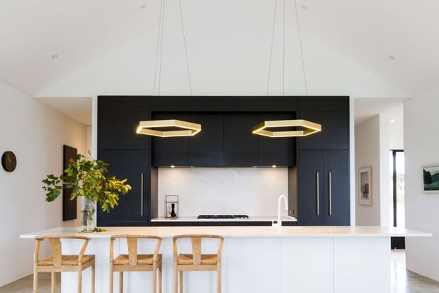Bright contemporary kitchen with a large white island and three woven timber bar stools, facing a wall of matte black cabinetry. A pale stone or composite benchtop, light backsplash, integrated cooktop, and white gooseneck tap are framed by two geometric brass-toned pendant lights. The room has a high white ceiling and opens toward a bright hallway or living area.