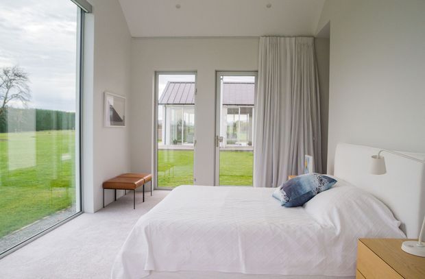Bright, minimalist bedroom with a low-profile white bed, white bedding, light grey walls, pale carpet, timber bedside table, and a small upholstered bench. Floor-to-ceiling glazing and tall glazed doors open views to a green lawn and adjacent pavilion-like structure, while full-height curtains provide privacy. The room has a clean, airy, restrained contemporary feel.